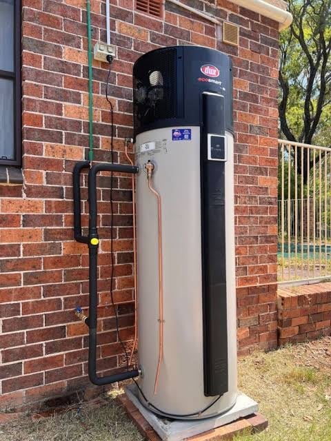 Black and white heat pump water heater installed against a brick house wall with copper piping