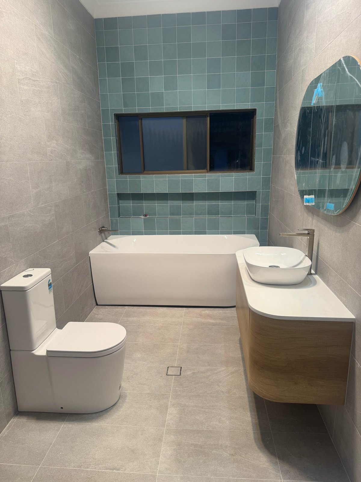 Modern bathroom with white toilet, oval bathtub, wooden vanity, round mirror, and teal tile accent wall with built-in niche